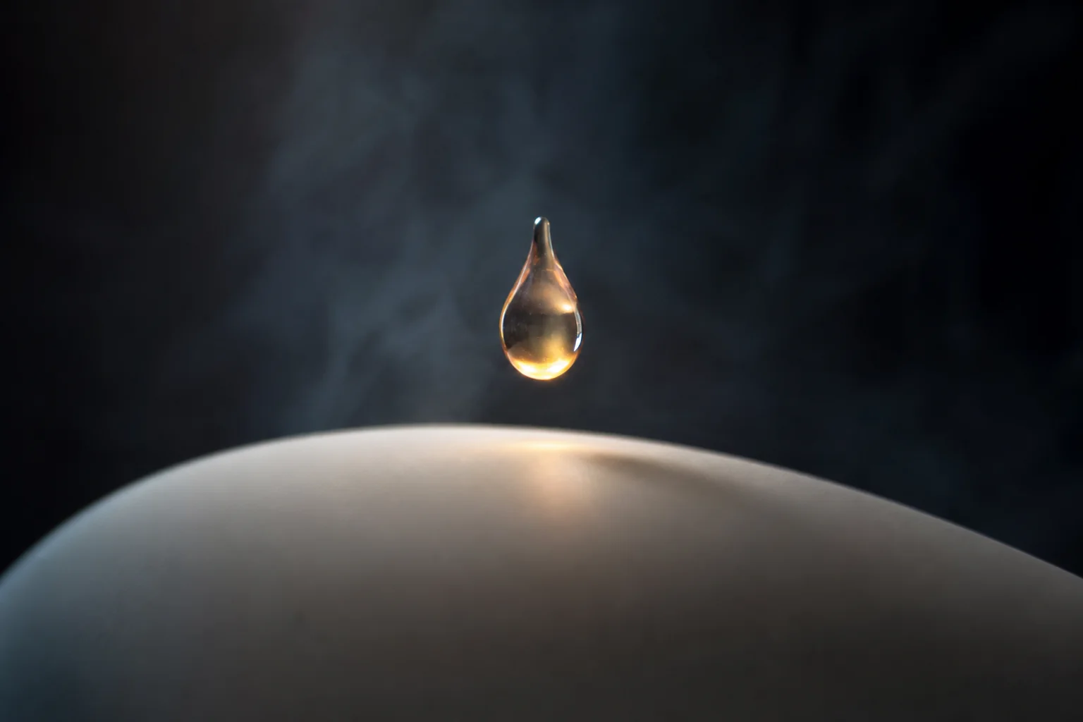 A glowing water droplet hangs above a smooth curved surface against a dark background.